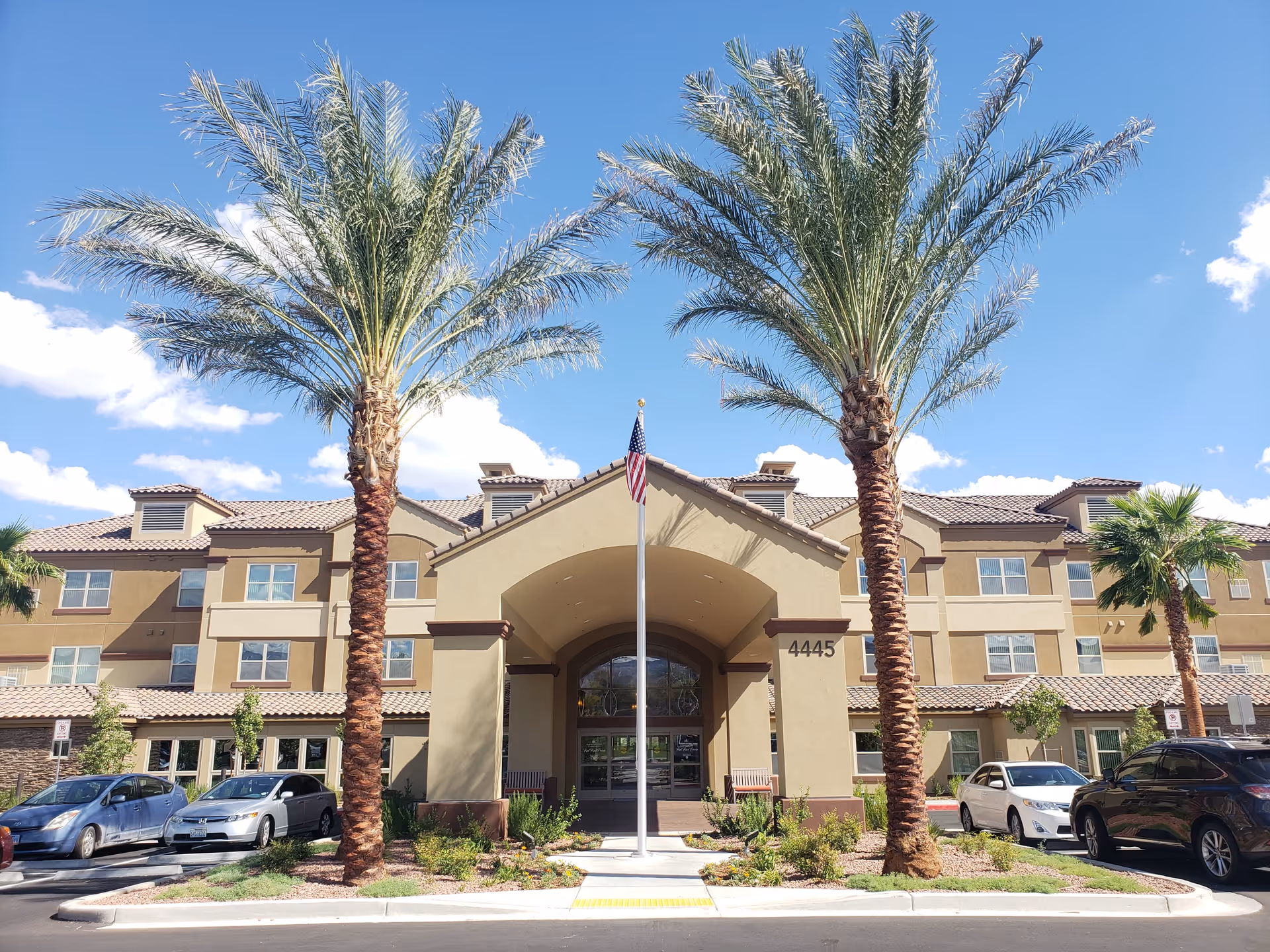 Front exterior view of Red Rock Pointe Retirement Community building with a central entrance, two tall palm trees, an American flag on a flagpole, and several parked cars under a clear blue sky.