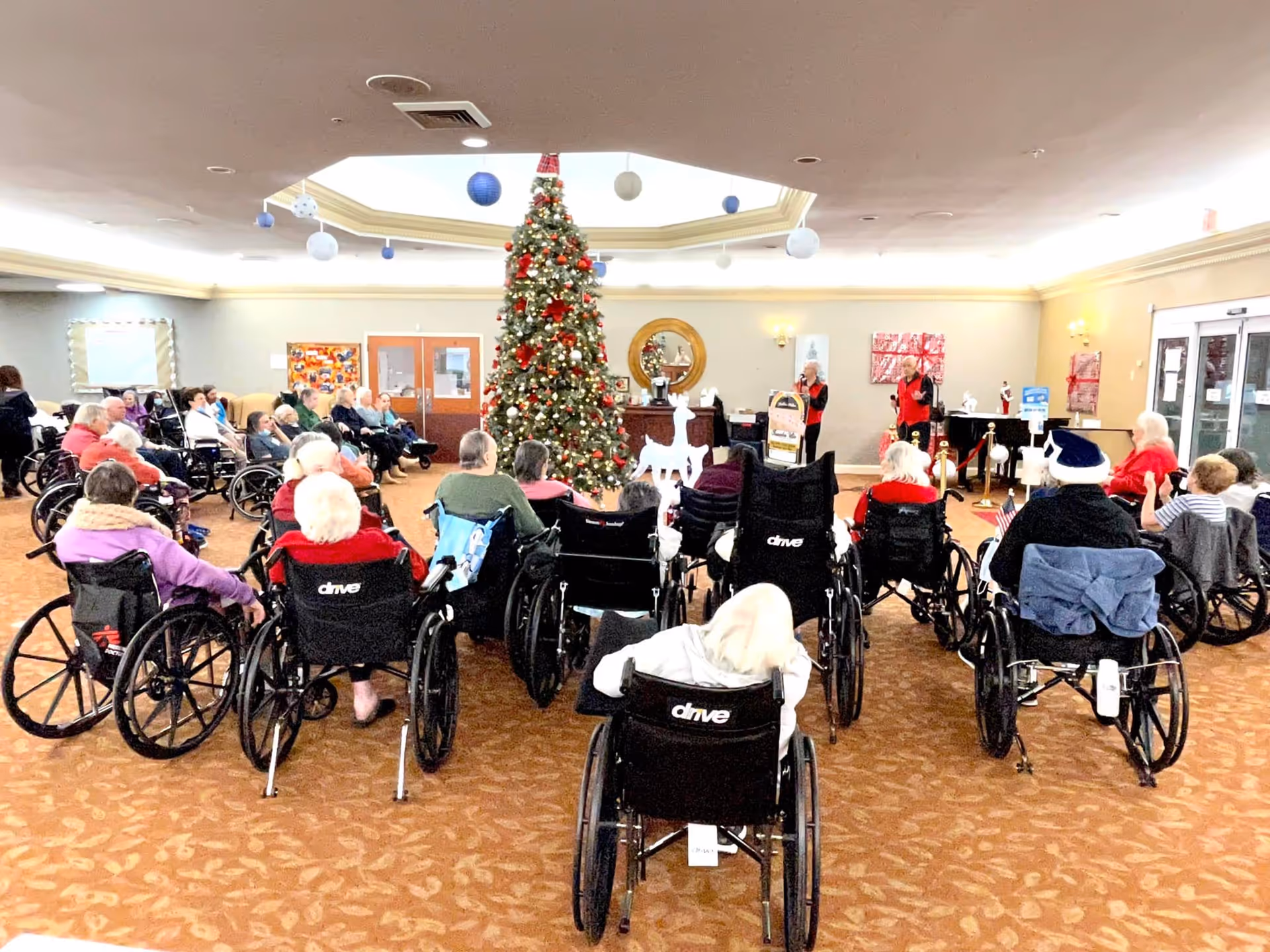 A group of elderly people, many in wheelchairs, seated in a semi-circle facing two performers in red vests in a decorated common area with a large Christmas tree and festive decorations.