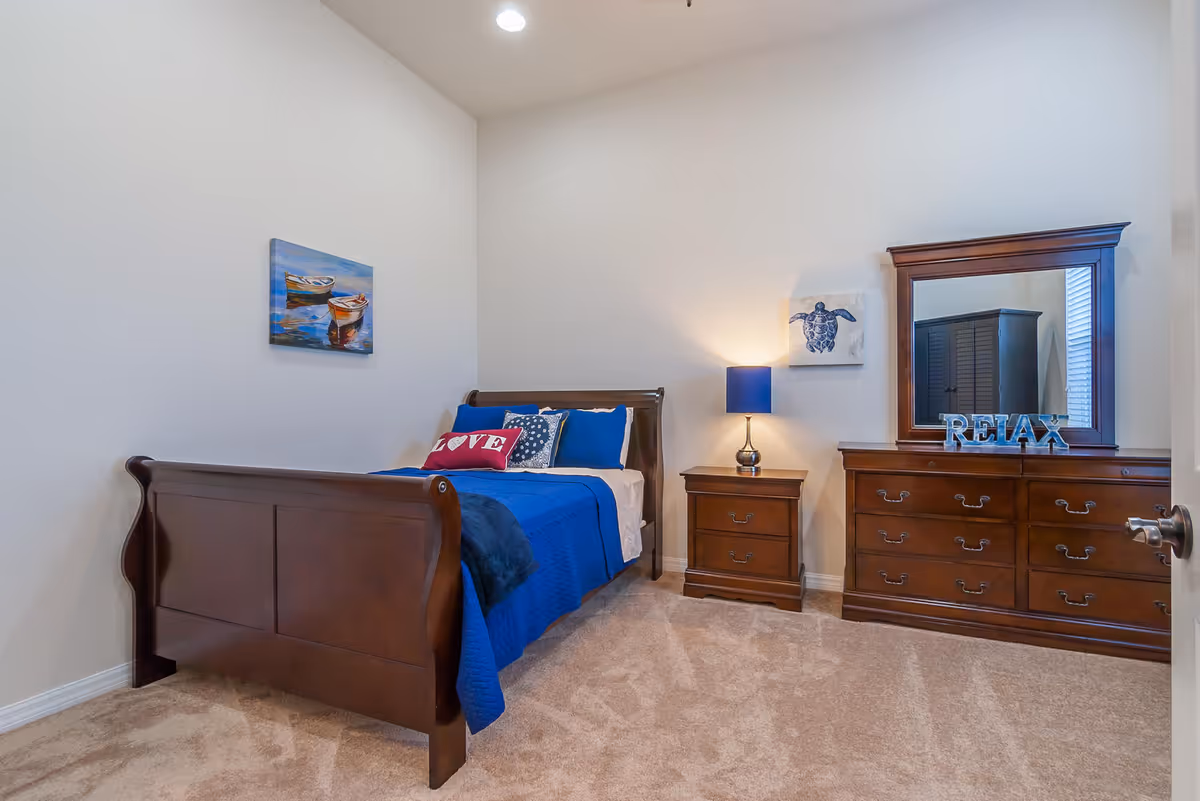 A bedroom with a dark wooden bed frame and blue bedding, including pillows with the word 'LOVE' and decorative patterns. There is a matching wooden nightstand with a blue lamp and a wooden dresser with a large mirror. On the dresser, there is a decorative sign that says 'RELAX'. The walls are light-colored with two paintings, one of boats on water and another of a turtle. The floor is carpeted in a light beige color.