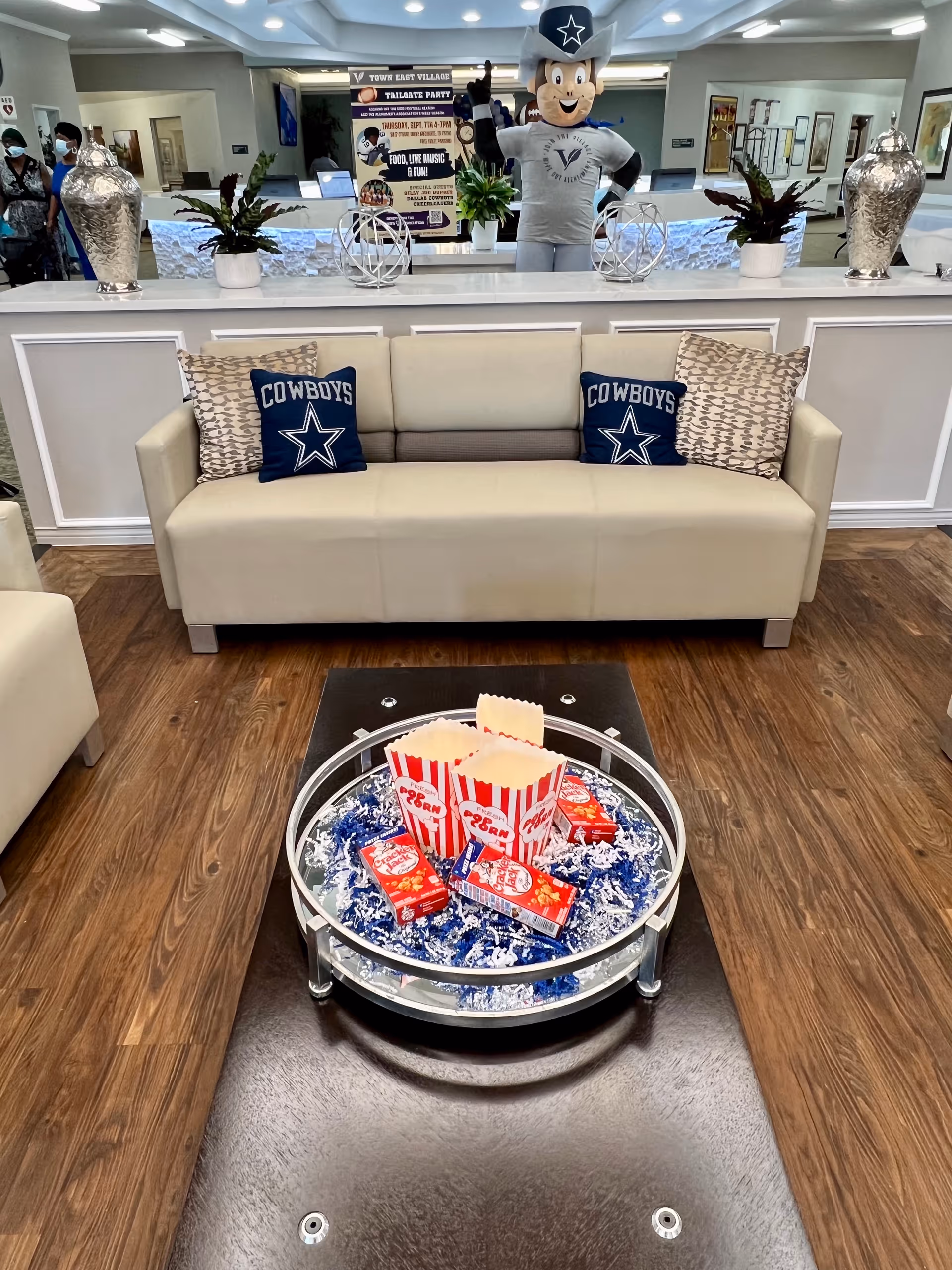 Lobby seating area with a beige sofa featuring Cowboys pillows, a coffee-table display of popcorn boxes and snacks, and a mascot and reception desk in the background.