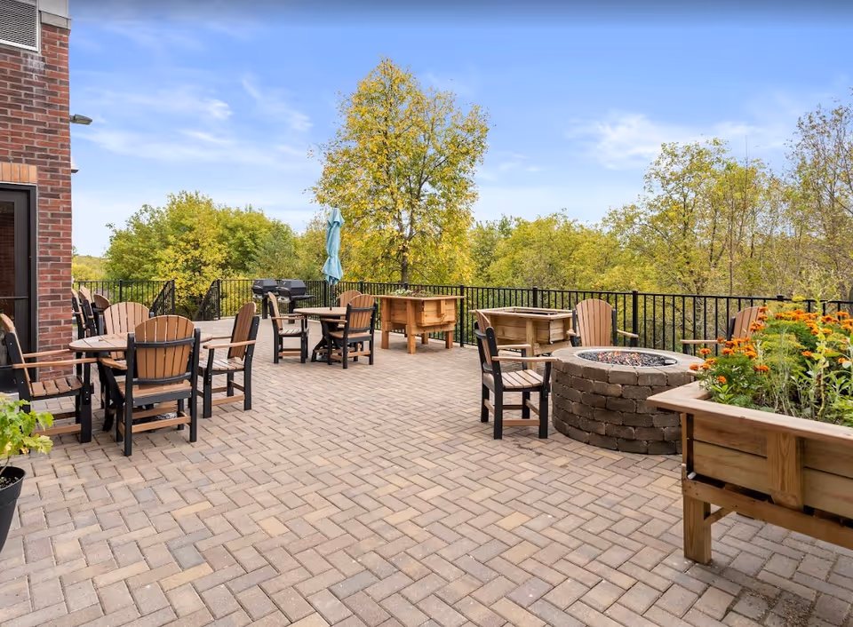 Outdoor patio area with brick flooring, several wooden tables and chairs, a fire pit, raised garden beds with flowers, and a grill. The patio is surrounded by a black metal railing and overlooks green trees under a blue sky.