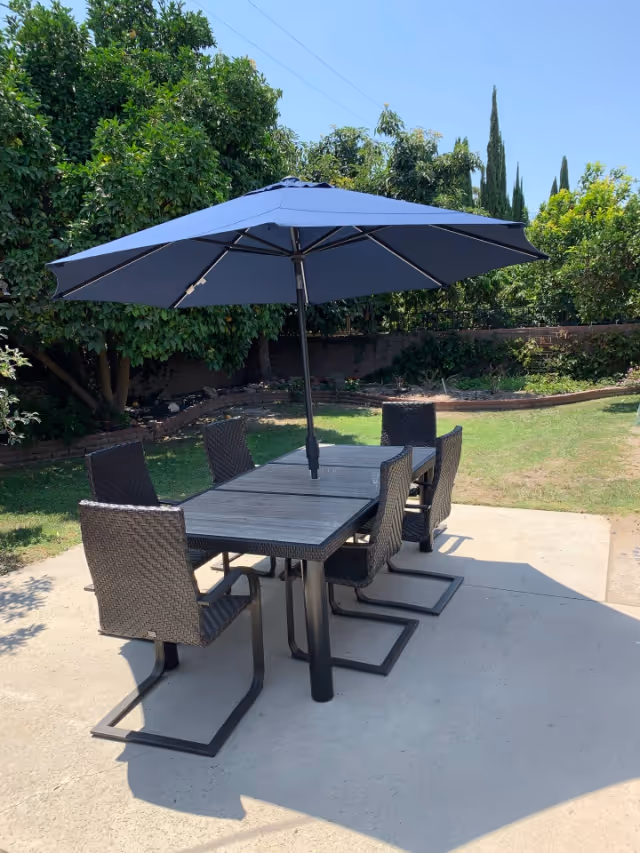 Outdoor patio area with a rectangular table and six woven chairs arranged around it. A large blue umbrella is open above the table providing shade. The patio is surrounded by green grass, trees, and shrubs under a clear blue sky.