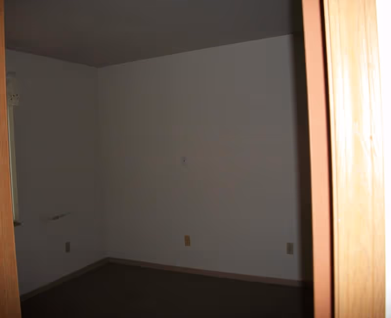 Empty room with beige walls, brown carpet, and wooden door frame. The room appears dimly lit with no furniture or decorations.