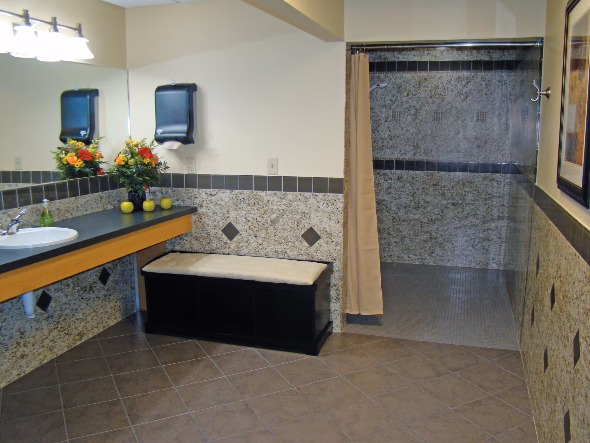 Accessible bathroom with a sink and large mirror, a cushioned bench, and a tiled walk-in shower with a curtain.