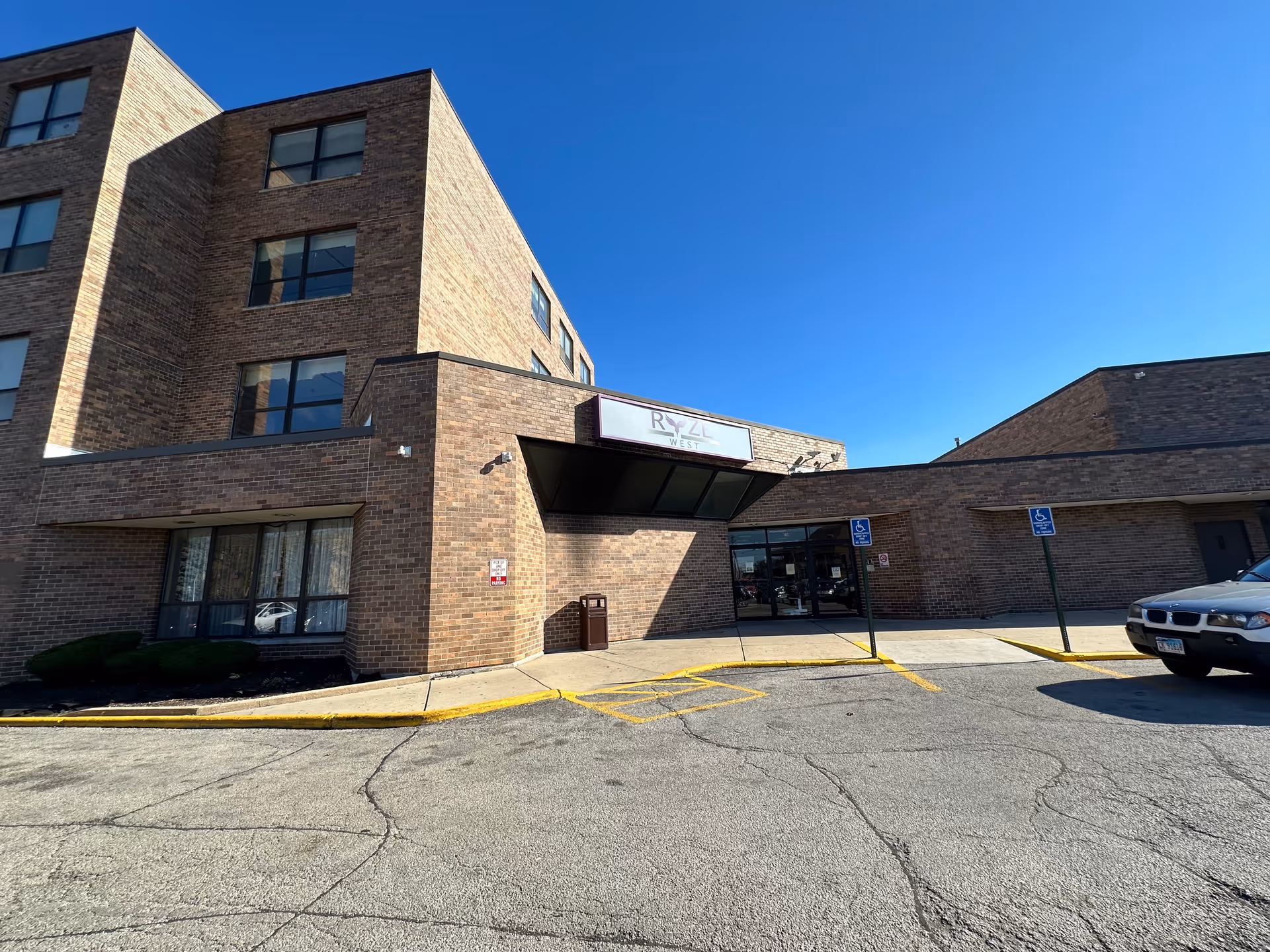 Front entrance of a brick multi-story building with a canopy and a sign reading 'RYZE West' under a clear blue sky.