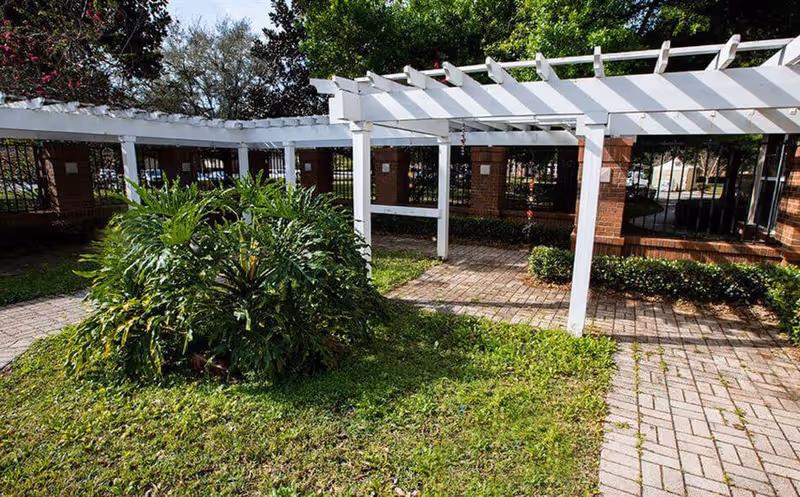 Outdoor courtyard featuring a white pergola over brick columns, paved walkways, and green shrubs.