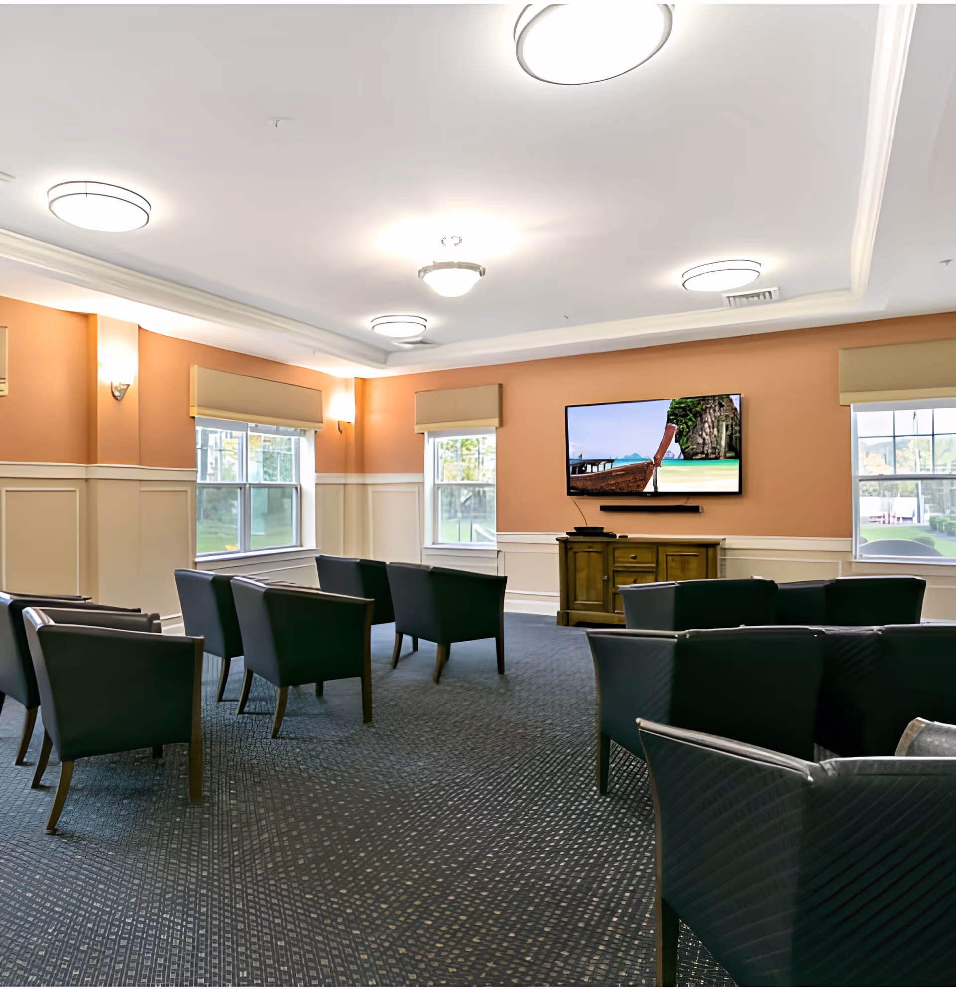 A well-lit common area with multiple black armchairs arranged in rows facing a wall-mounted flat-screen TV displaying a scenic beach image. The room has peach-colored walls with white wainscoting, three windows with beige valances, and a wooden cabinet beneath the TV. The ceiling features several round light fixtures.