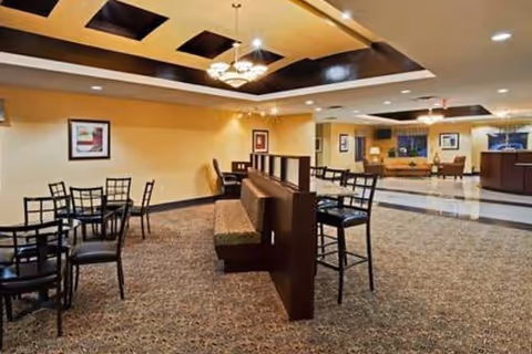 Interior view of a senior living facility common area with carpeted floor, several black metal chairs and tables, a wooden partition with cushioned bench seating, warm yellow walls, framed artwork, and a ceiling with recessed lighting and decorative panels.