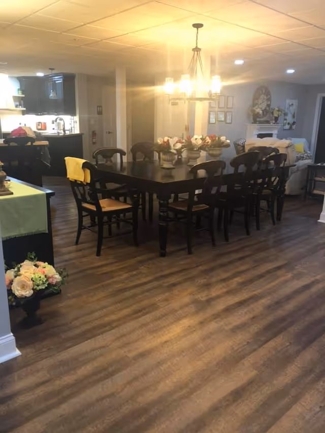 Interior view of a dining area in Magnolia Assisted Living featuring a large dark wooden dining table with eight chairs. The table is decorated with flower arrangements. A chandelier with multiple lights hangs above the table. In the background, there is a kitchen area with dark cabinets and a living room with a fireplace, wall clock, and framed pictures. The floor is wooden and there are additional floral decorations near the entrance.