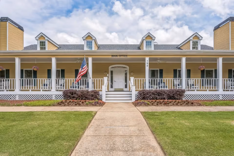 Front exterior view of a yellow building with white trim and a porch with white railings. There are several windows with dark shutters, hanging flower baskets, and an American flag near the entrance. The building has a sign above the door that reads 'Main Office' and the number 150 is displayed on a column. A concrete walkway leads up to the entrance, and there is green grass and landscaping in front.