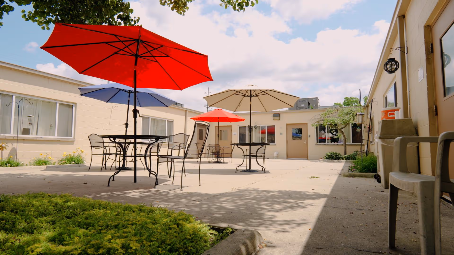 Outdoor courtyard area with several metal tables and chairs, each shaded by large colorful umbrellas in red, blue, and beige. The courtyard is surrounded by single-story building walls with windows and doors, some greenery, and a partly cloudy sky overhead.