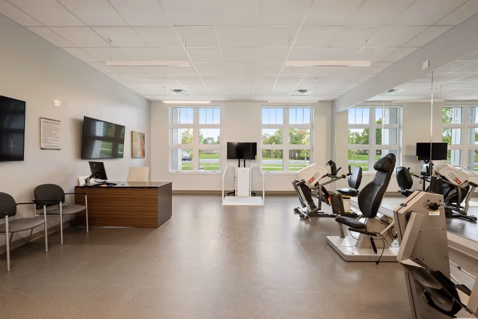 A bright and spacious exercise room with large windows letting in natural light. The room features exercise equipment including stationary bikes and a recumbent bike, a desk with a computer, chairs along the wall, and a large mirror covering one side of the room.