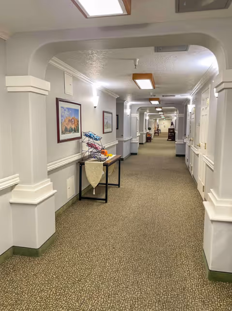 A long, carpeted hallway in a senior living facility with white walls and ceiling. The hallway features decorative arches, wall-mounted lights, framed artwork, and a small table with a wire rack and some items on it. Doors line the right side of the hallway, and the corridor extends into the distance with more furniture and lighting visible.
