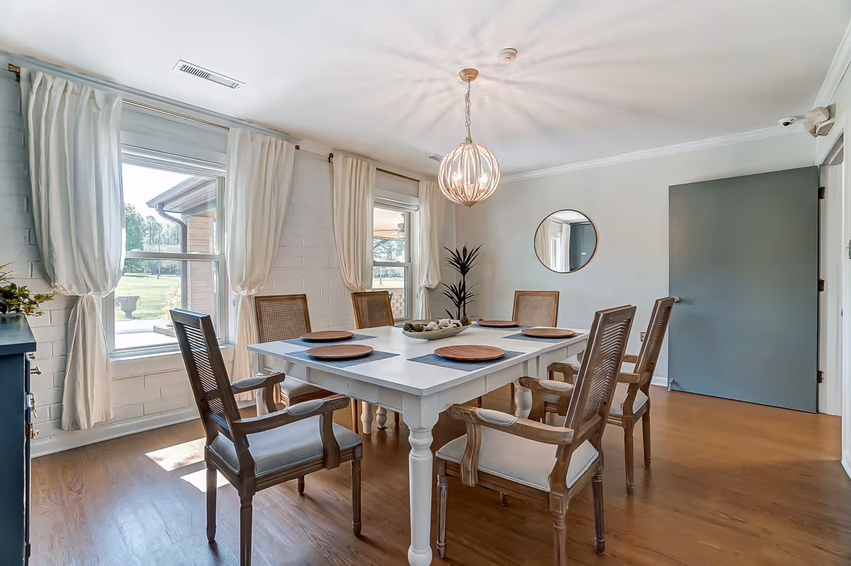 A bright dining room with a white rectangular table set for six with wooden chairs. The room has two large windows with white curtains, a round mirror on the wall, a modern spherical chandelier hanging from the ceiling, and a potted plant in the corner. The floor is wooden, and there is a closed gray door on the right side.