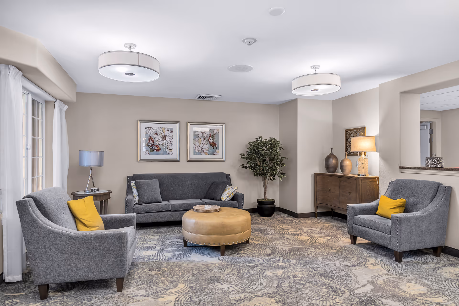 A furnished lounge with gray sofa and armchairs, a round leather ottoman, lamps, artwork, and a potted plant.