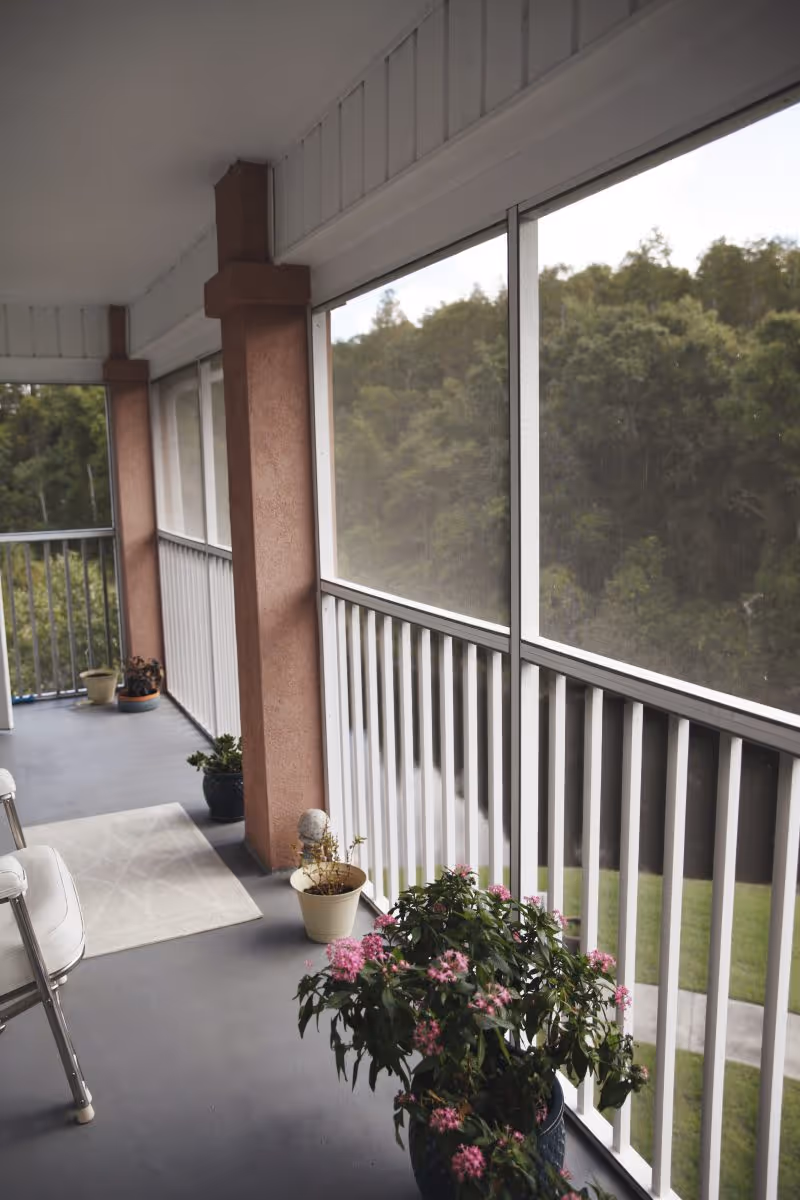 Screened balcony with potted plants, seating, and a railing overlooking trees.