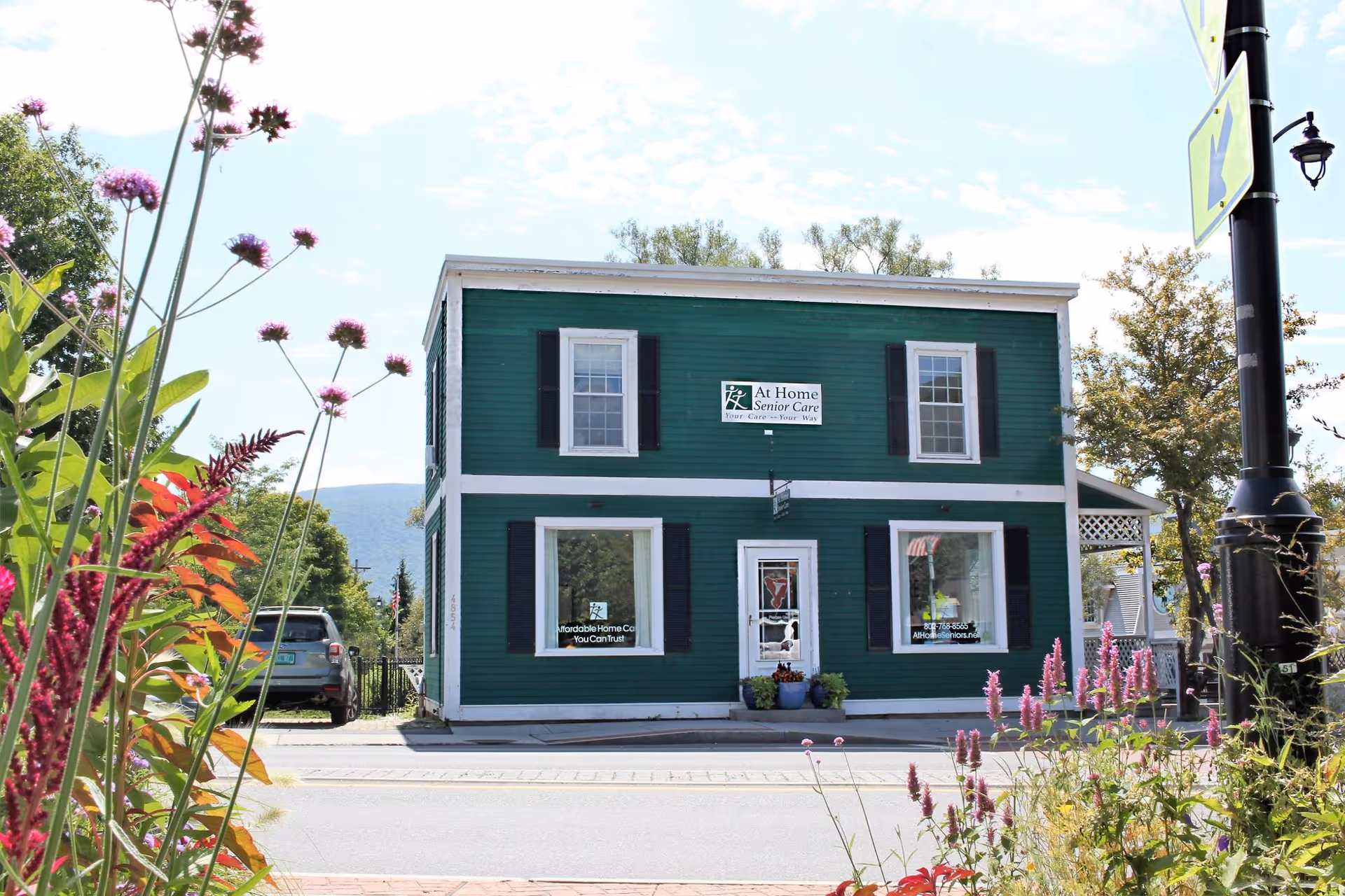 Two-story green 'At Home Senior Care' building facing the street with flowering plants in the foreground.