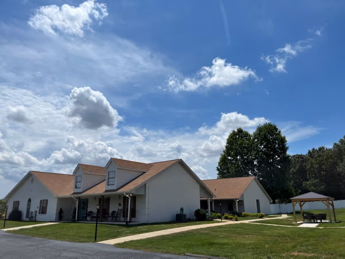Exterior view of a single-story senior living facility building with a brown roof and white walls under a partly cloudy blue sky. There is a small covered porch with chairs, a paved walkway, green lawn, trees, and a wooden gazebo with a table and chairs on the right side.