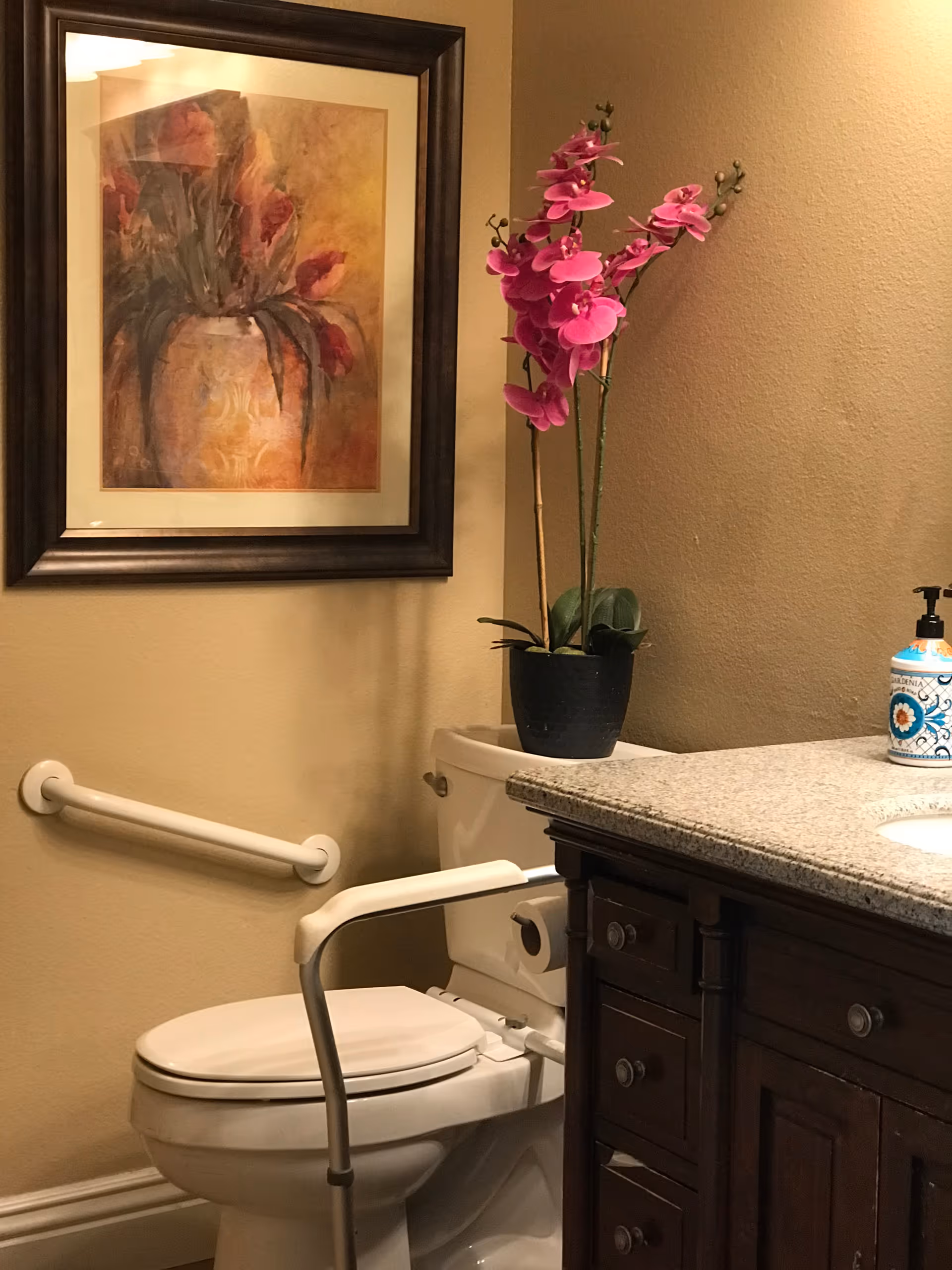 A bathroom with a white toilet equipped with a safety grab bar and a support arm. Above the toilet is a framed painting of flowers in a vase. To the right, there is a granite countertop with a dark wooden cabinet underneath, a potted pink orchid, and a decorative soap dispenser.