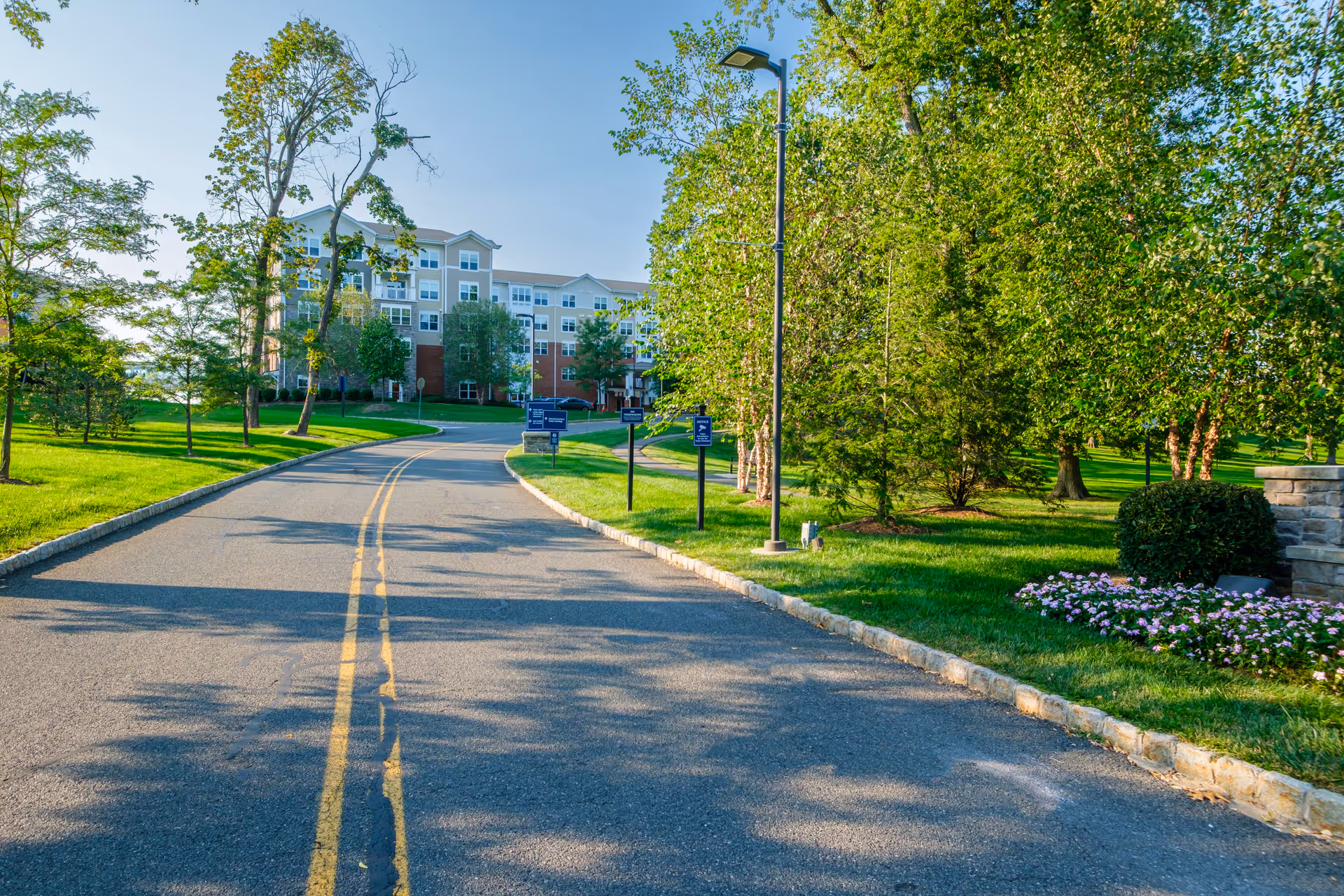 A paved road with double yellow lines curves gently through a landscaped area with green grass, trees, and flower beds. On the left side, there are several trees and a large multi-story residential building in the background. On the right side, there are more trees, a street lamp, and a stone wall with flowers. The sky is clear and blue.