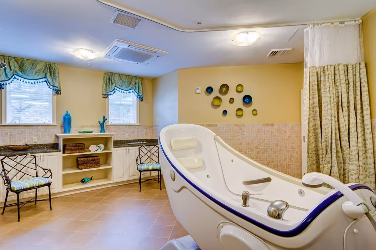 A spacious bathroom with a large white and blue therapeutic bathtub in the foreground. The room has beige tiled floors and walls with a decorative tile border. Two windows with green valances let in natural light. There are two chairs with striped cushions and a built-in cabinet with shelves holding towels and decorative items. A curtain is partially drawn on the right side, and decorative plates are mounted on the wall.