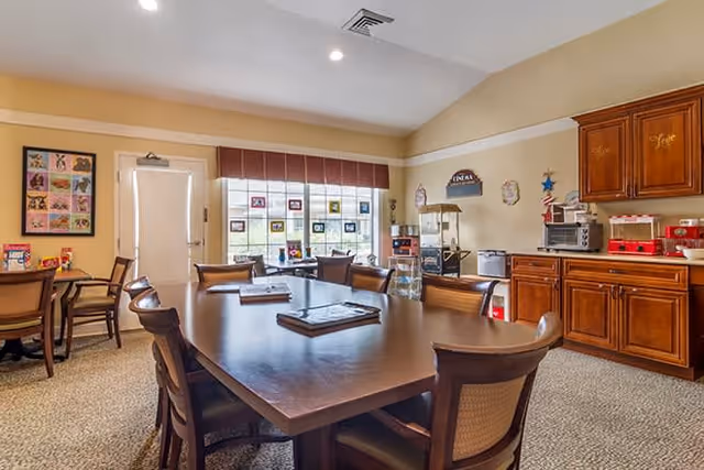 A well-lit room with a large wooden table surrounded by chairs, a window with blinds letting in natural light, wooden cabinets along one wall, and various kitchen appliances including a microwave, popcorn machine, and coffee maker.