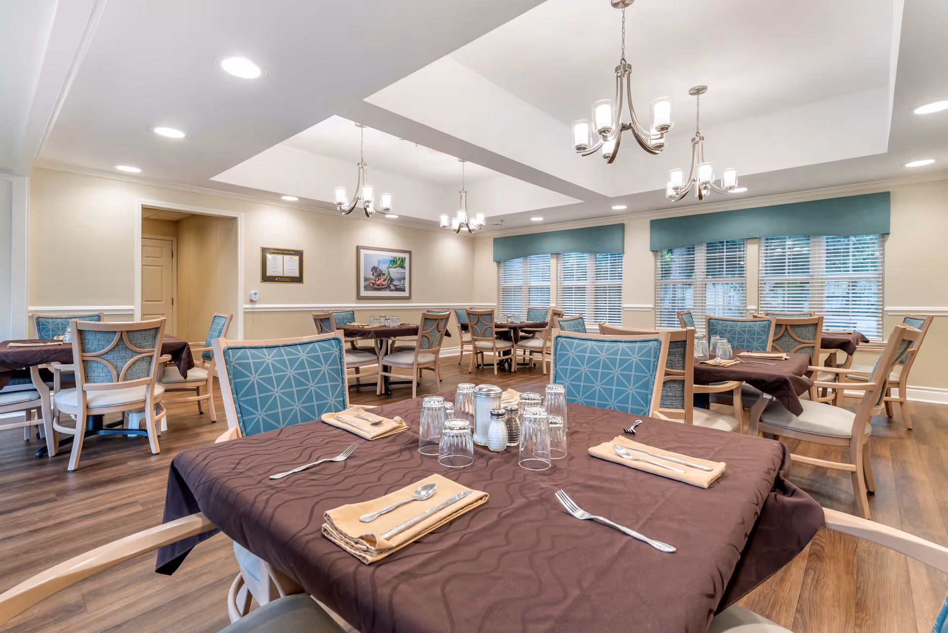 A dining room in a senior living facility with multiple tables covered in brown tablecloths, each set with utensils, napkins, and upside-down drinking glasses. The chairs have light wood frames with blue patterned upholstery. The room features wooden flooring, large windows with blue valances, and modern chandeliers hanging from a white ceiling with recessed lighting.