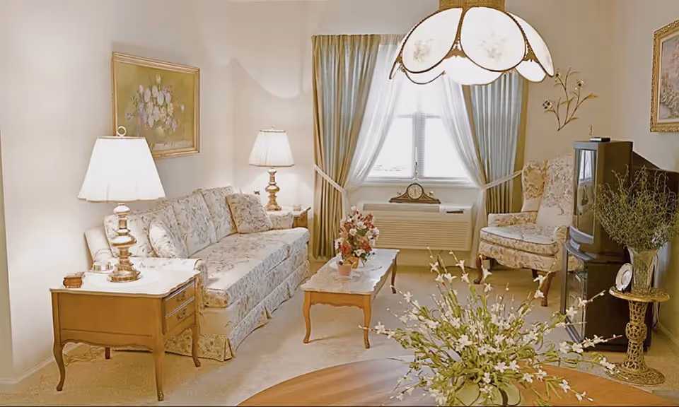 Bright, traditionally furnished living room with a floral sofa and armchair, coffee table, lamps, window with drapes, and a TV.