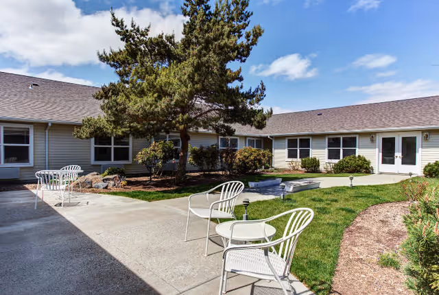Outdoor courtyard area at Carriage Place Memory Care with white metal chairs and tables on a concrete patio, a small grassy area with landscaping including a tree and bushes, and a single-story building with multiple windows and doors surrounding the courtyard under a partly cloudy sky.