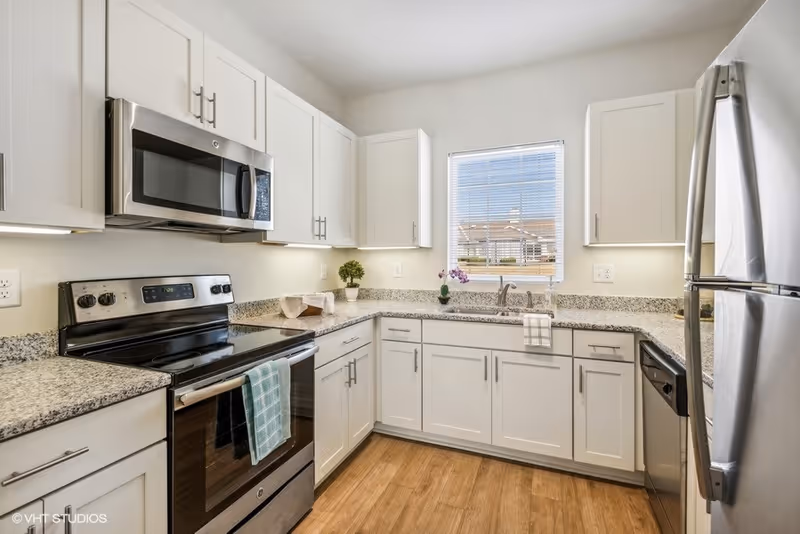 Modern kitchen with white cabinets, granite countertops, stainless steel appliances including a stove, microwave, refrigerator, and dishwasher. A window with blinds is above the sink, and there are small decorative plants on the countertop. The floor is wood-style.