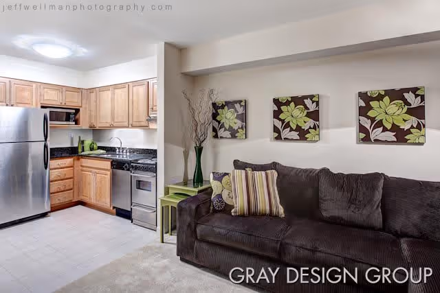 Interior view of a living space featuring a small kitchen area with wooden cabinets, stainless steel refrigerator, stove, and dishwasher. Adjacent to the kitchen is a dark brown sofa with striped and floral pillows, a green side table with a tall vase holding decorative branches, and three floral wall art pieces above the sofa.