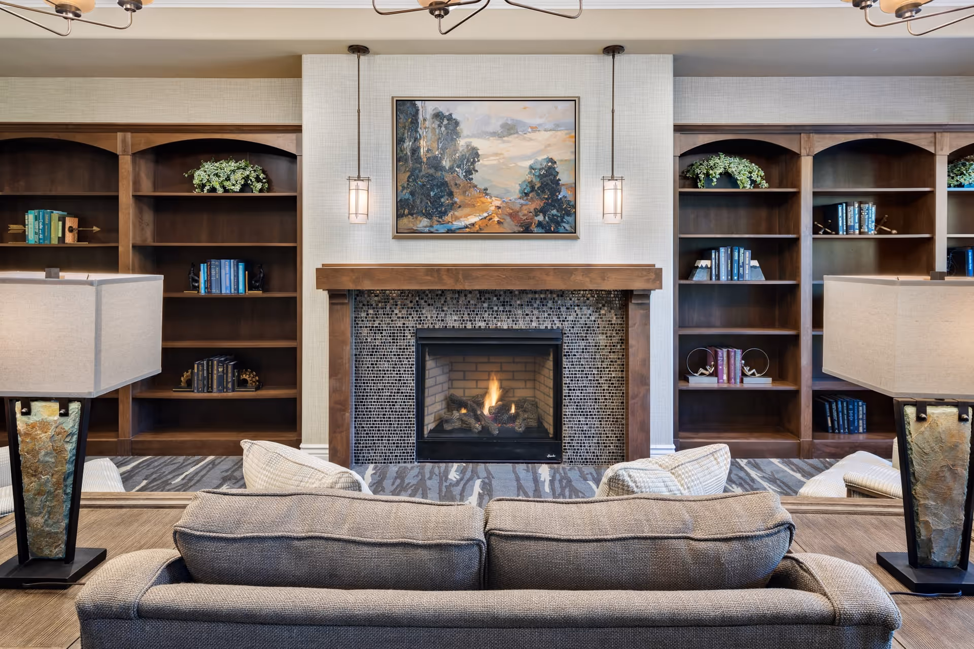 Cozy common room with a sofa facing a lit fireplace flanked by built-in wooden bookshelves, lamps, and artwork above the mantel.