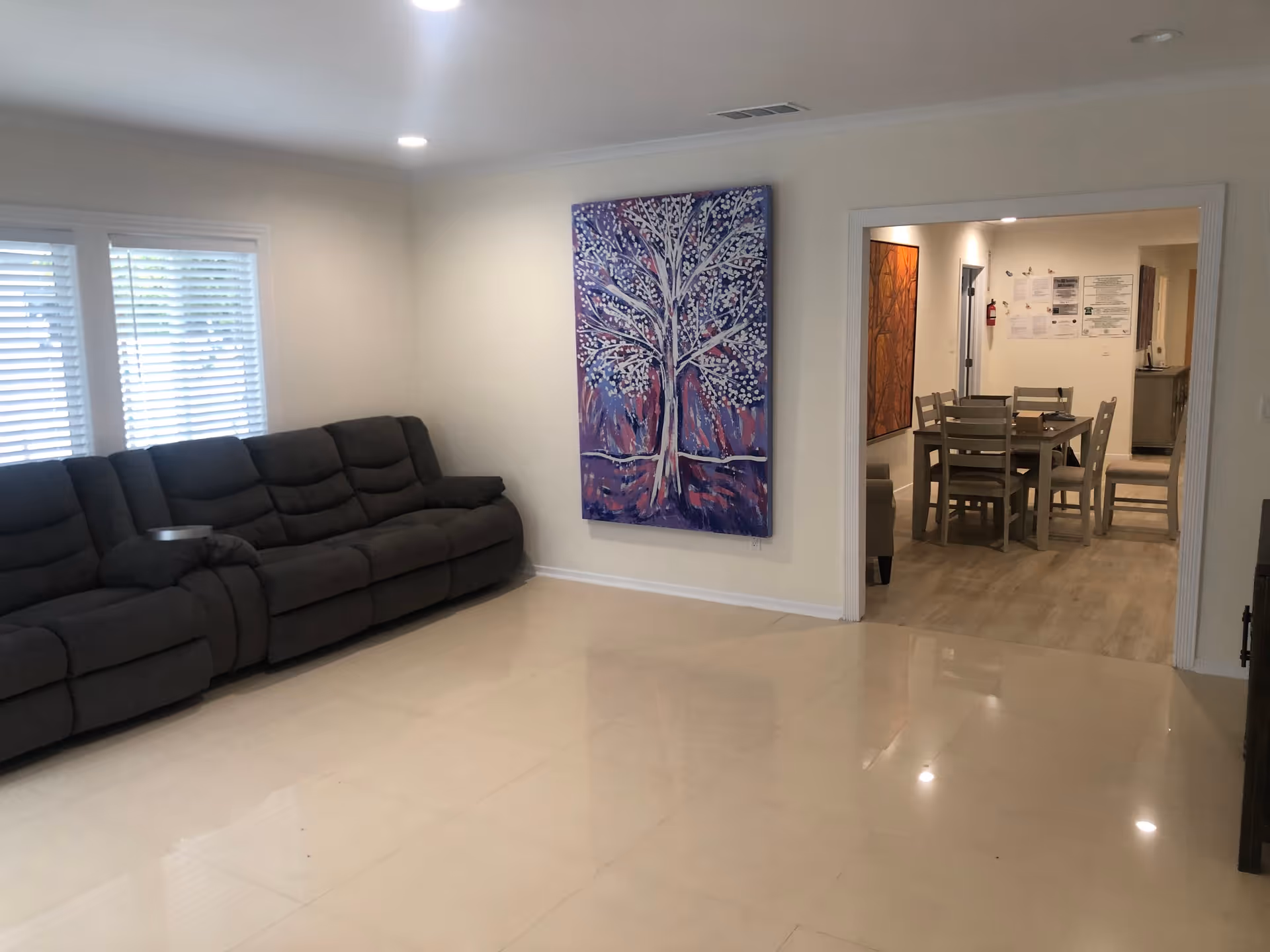 A living room with a large dark gray sectional sofa along the left wall, two windows with white blinds, and a large colorful painting of a tree on the wall. Through an open doorway, a dining area with a wooden table and six chairs is visible, along with some wall decorations and a fire extinguisher.