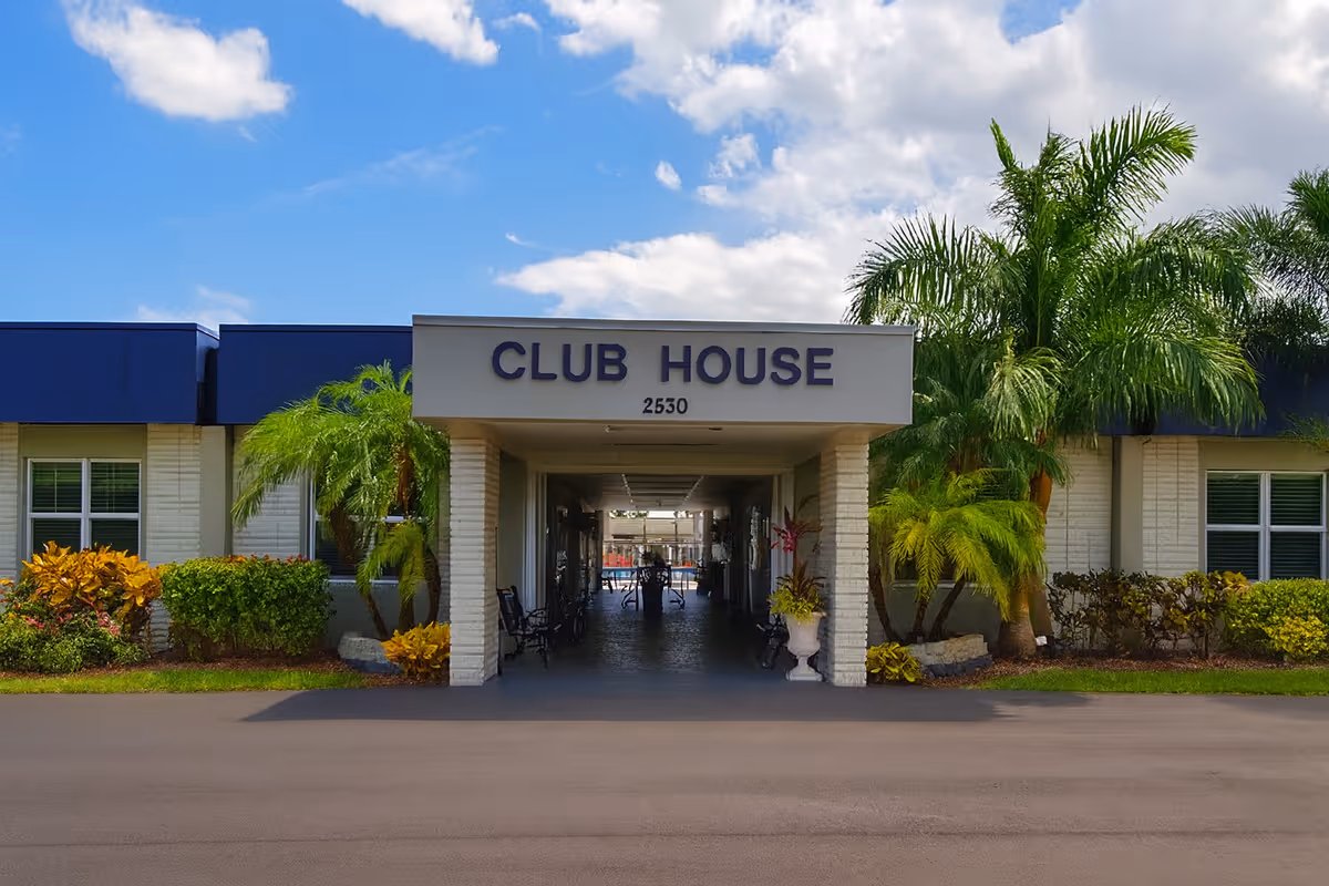Front entrance of a low-rise clubhouse labeled 'CLUB HOUSE' with palm trees and a covered walkway.