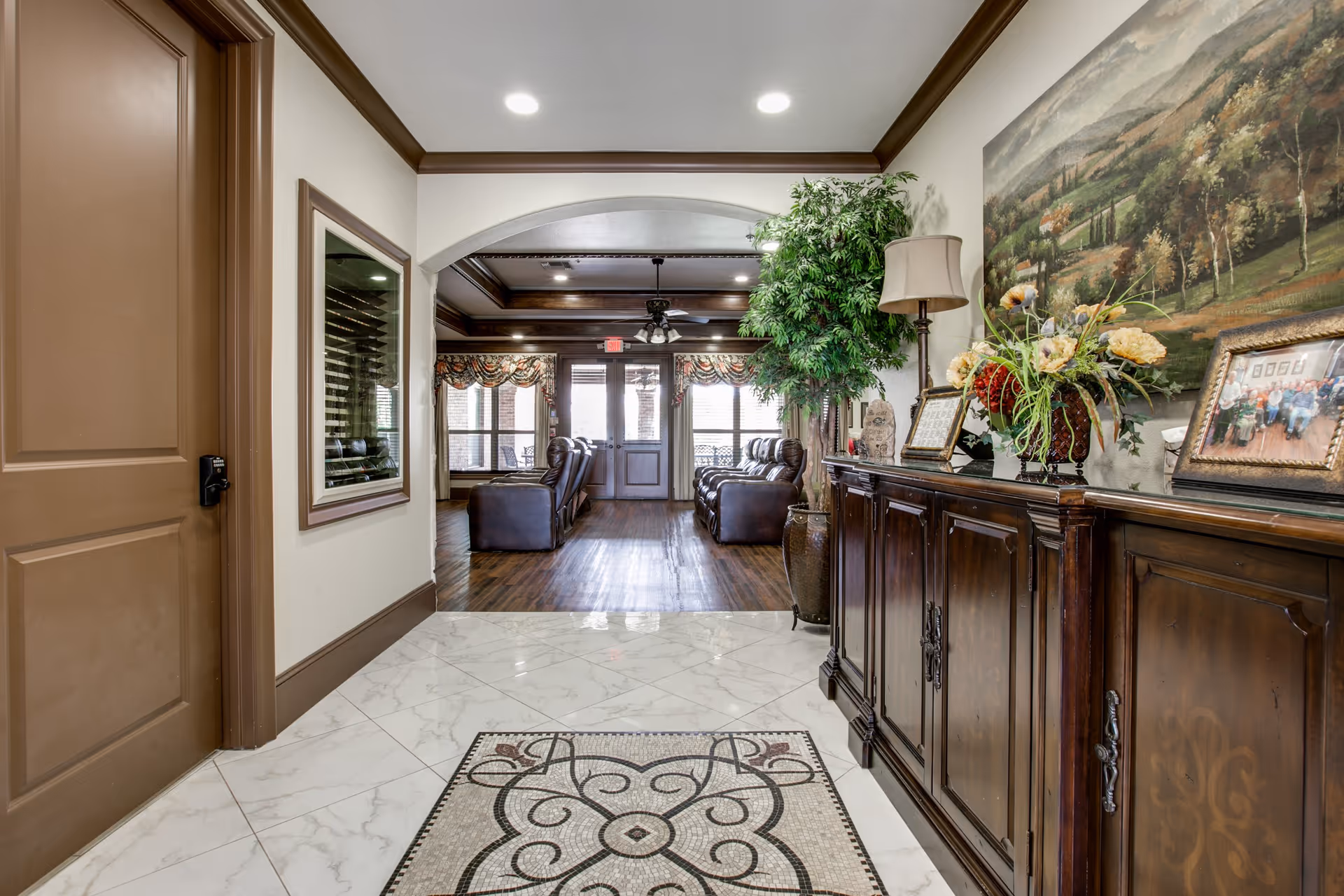 Interior view of a senior living facility hallway leading into a common sitting area with leather armchairs. The hallway has a decorative tile floor with a patterned rug, a wooden sideboard with floral arrangements and framed photos, a large painting on the wall, and a tall potted plant. The sitting area beyond has wooden flooring, ceiling fans, and large windows with patterned valances.