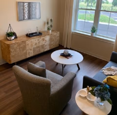 A cozy living room with a gray armchair and a dark gray sofa with a yellow pillow. There are two round coffee tables, one with a decorative item and the other with a small plant and two white cups. A wooden sideboard with decorative items and a framed artwork hangs on the wall. Large windows let in natural light and show a view of greenery outside.