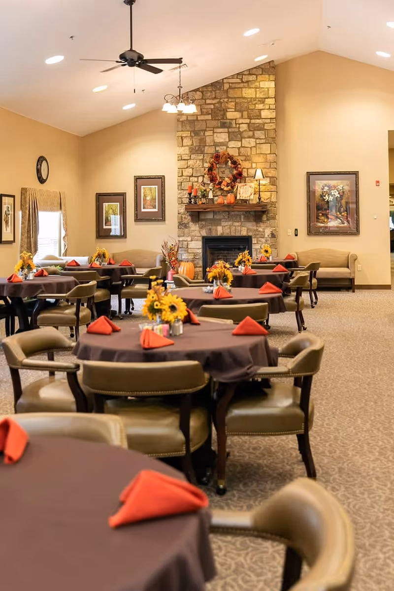 A warm and inviting dining room with round tables covered in brown tablecloths and set with red folded napkins. Each table has a centerpiece of sunflowers. The room features a stone fireplace decorated with autumn-themed items including a wreath, pumpkins, and candles. Comfortable chairs surround the tables, and framed artwork and a clock adorn the beige walls. The ceiling has recessed lighting and a ceiling fan.