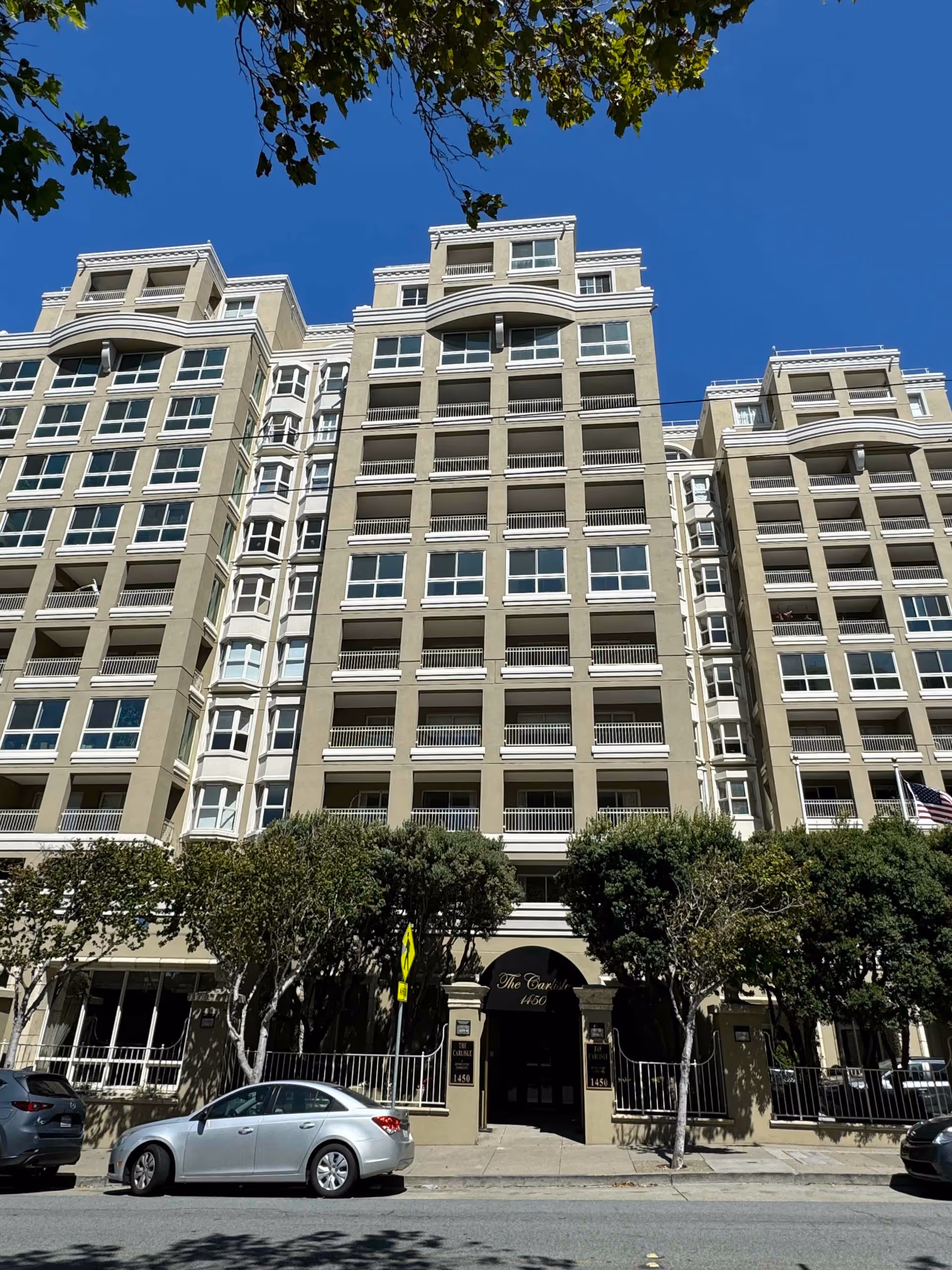 Exterior front view of The Carlisle, a multi-story residential building with balconies and numerous windows, surrounded by trees and parked cars on the street in front.