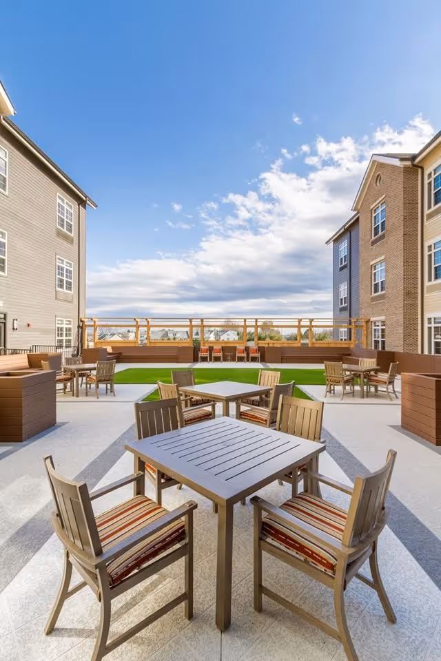 Outdoor patio area at Tribute at The Glen with multiple wooden tables and chairs featuring striped cushions, surrounded by multi-story residential buildings under a partly cloudy blue sky.