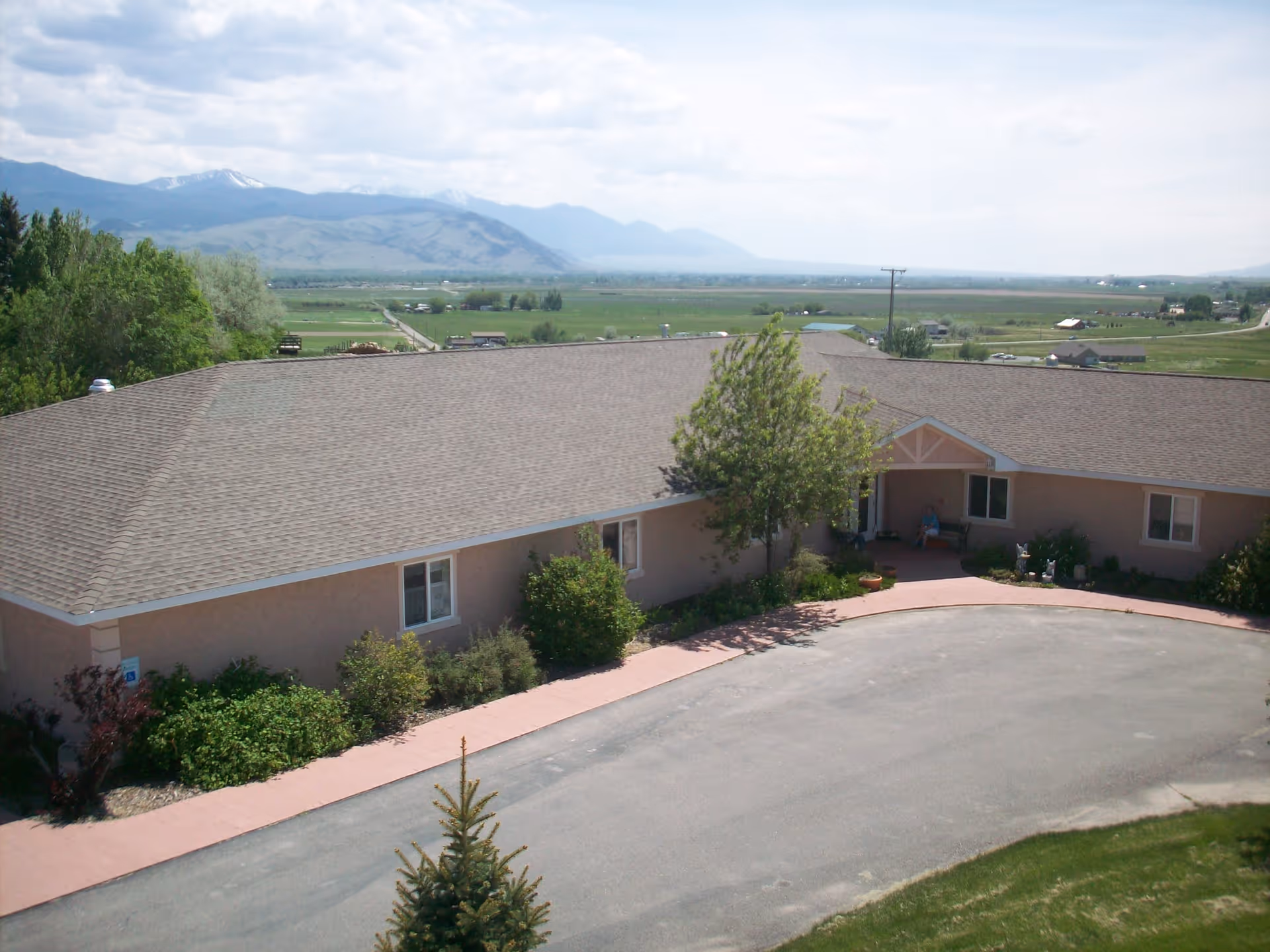 Exterior view of Meadowlark Manor, a single-story building with a sloped roof, surrounded by greenery and a paved driveway. In the background, there are expansive fields and mountains under a partly cloudy sky.