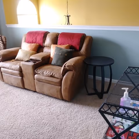 A cozy living room area with two brown leather recliner chairs, each with a red throw blanket and multiple cushions. There is a small round black side table between the chairs and a glass coffee table with a hand sanitizer bottle and magazines on it. The walls are painted in two tones, yellow on the upper half and blue on the lower half, separated by a white trim.