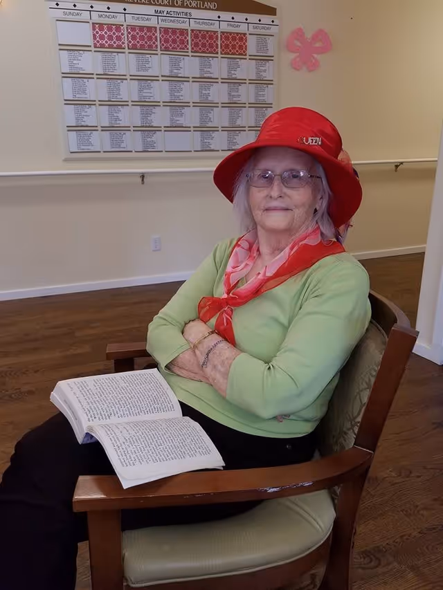 An elderly woman wearing a red hat and a green long-sleeve shirt sits in a wooden chair with a book open on her lap. She is indoors, with a large activity calendar on the wall behind her.