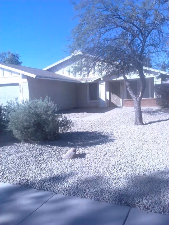 Front exterior view of a single-story house with a gravel yard, a tree, and some bushes under a clear blue sky.