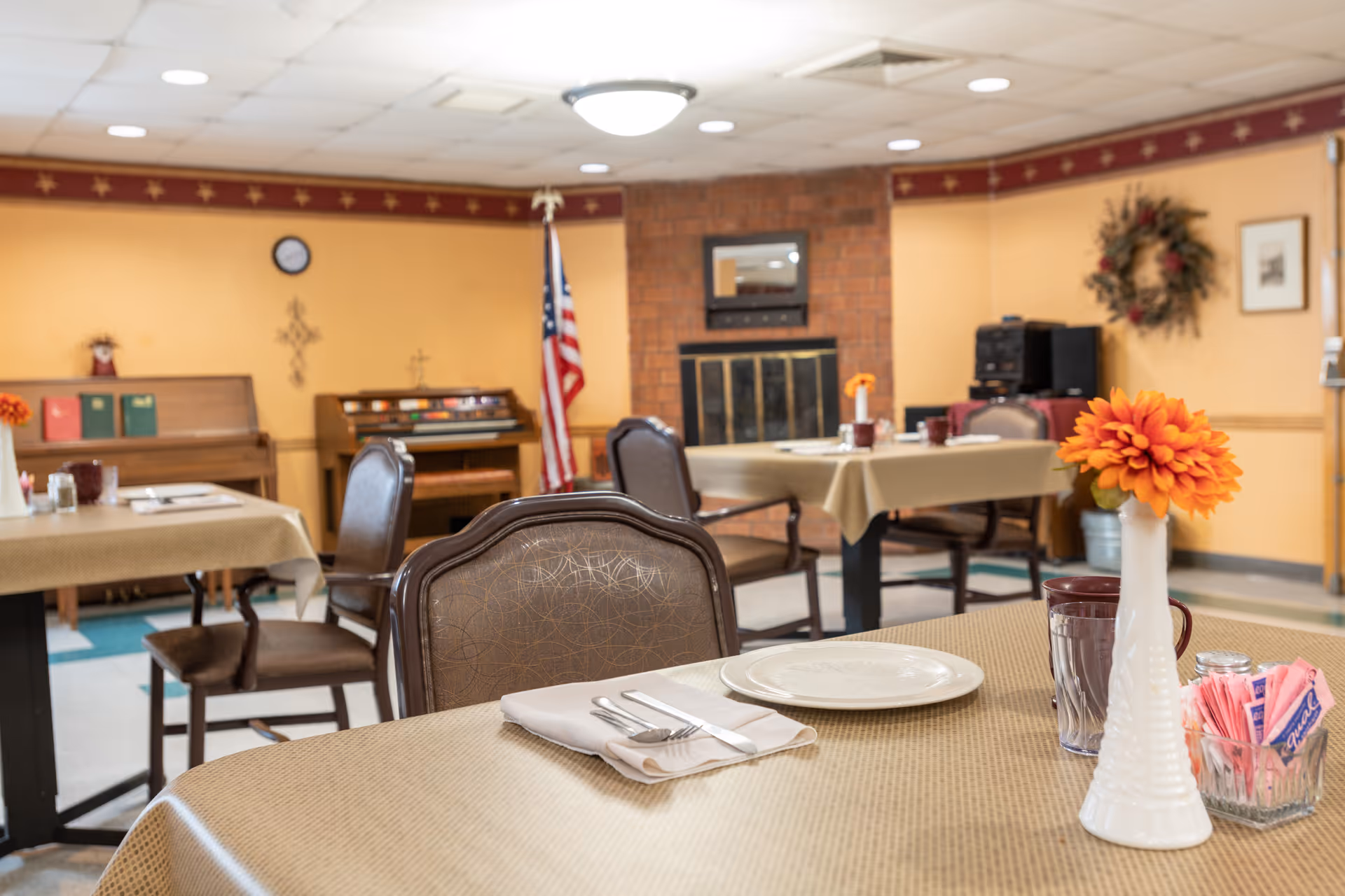 A dining room in a senior living facility with tables covered in beige tablecloths, set with plates, utensils, and cups. There are brown chairs around the tables, a vase with orange flowers, and a container with sugar packets. In the background, there is a piano, an American flag, a brick fireplace, and a wreath on the wall.