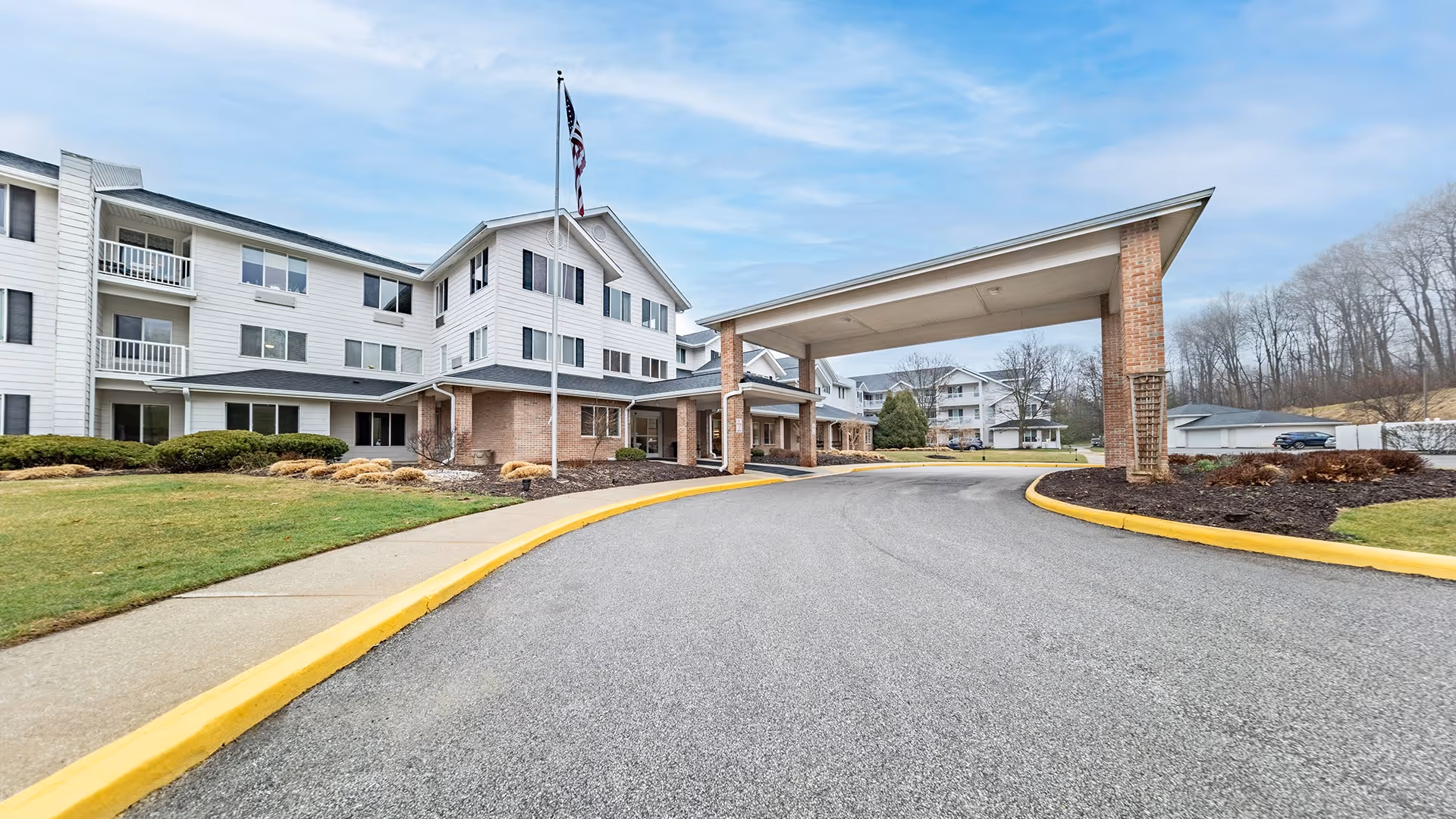 Exterior view of StoryPoint Strongsville senior living facility showing a large white multi-story building with a covered entrance driveway, an American flag on a flagpole, landscaped bushes, and a clear sky.