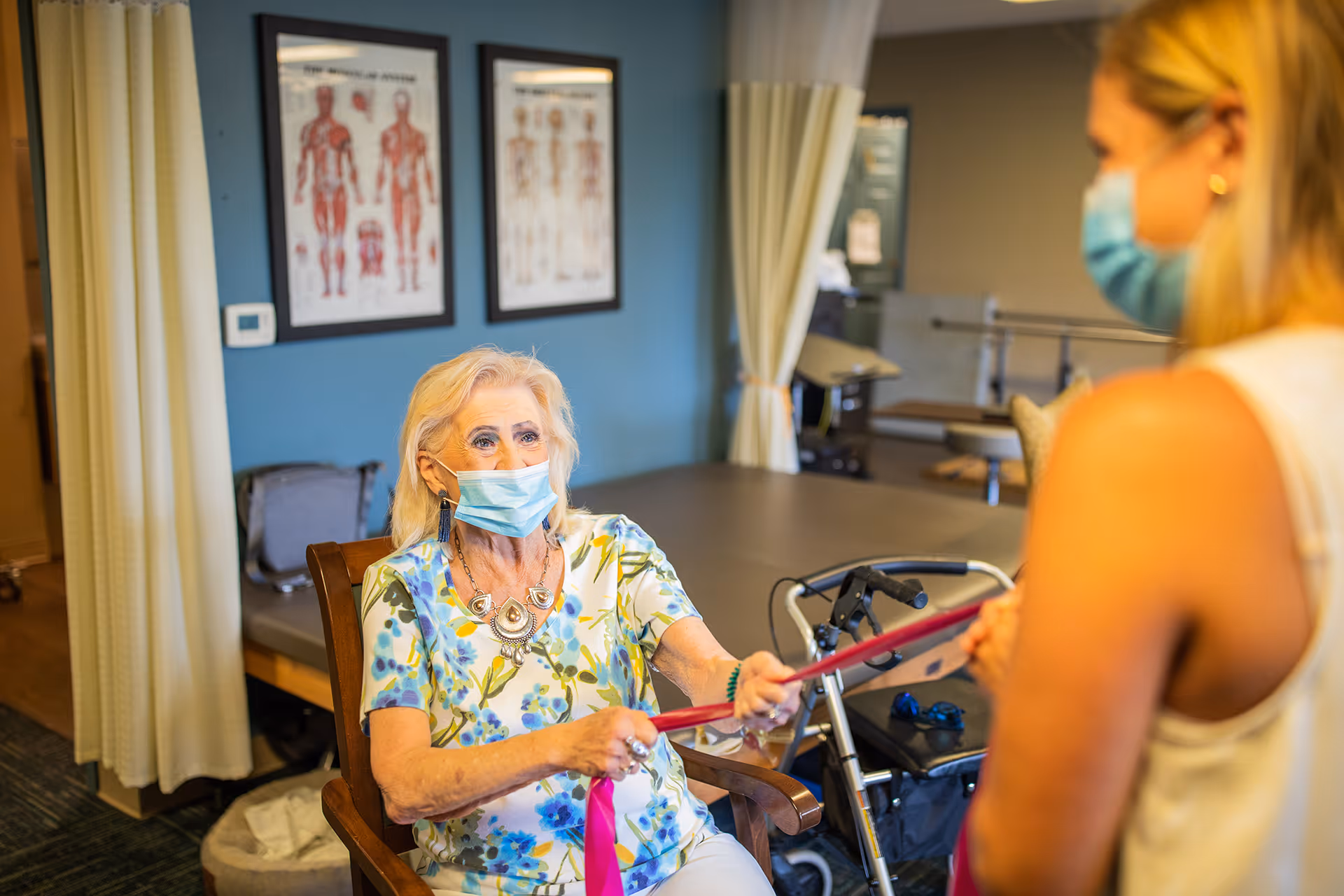 An elderly woman wearing a face mask is seated and holding a resistance band being pulled by a caregiver, who is also wearing a face mask. The setting appears to be a physical therapy or rehabilitation room with anatomical charts on the wall and medical equipment in the background.