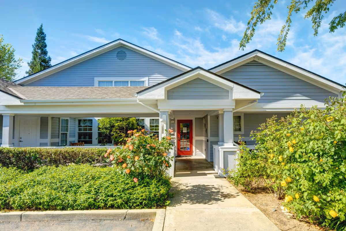 Exterior view of a single-story building with gray siding and a red door entrance, surrounded by green bushes and flowering plants under a blue sky with some clouds.