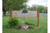Wooden sign for Willow Ridge Healthcare mounted on two posts, situated on a grassy area with a small landscaped garden in front, trees and a road visible in the background.
