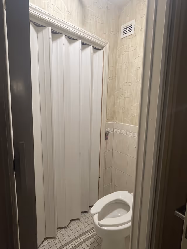 Small restroom featuring a toilet, a white folding door, tiled floor, and patterned wallpaper.