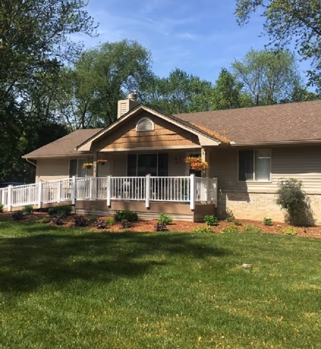 Single-story beige senior home with white railing porch, manicured lawn, and surrounding trees.